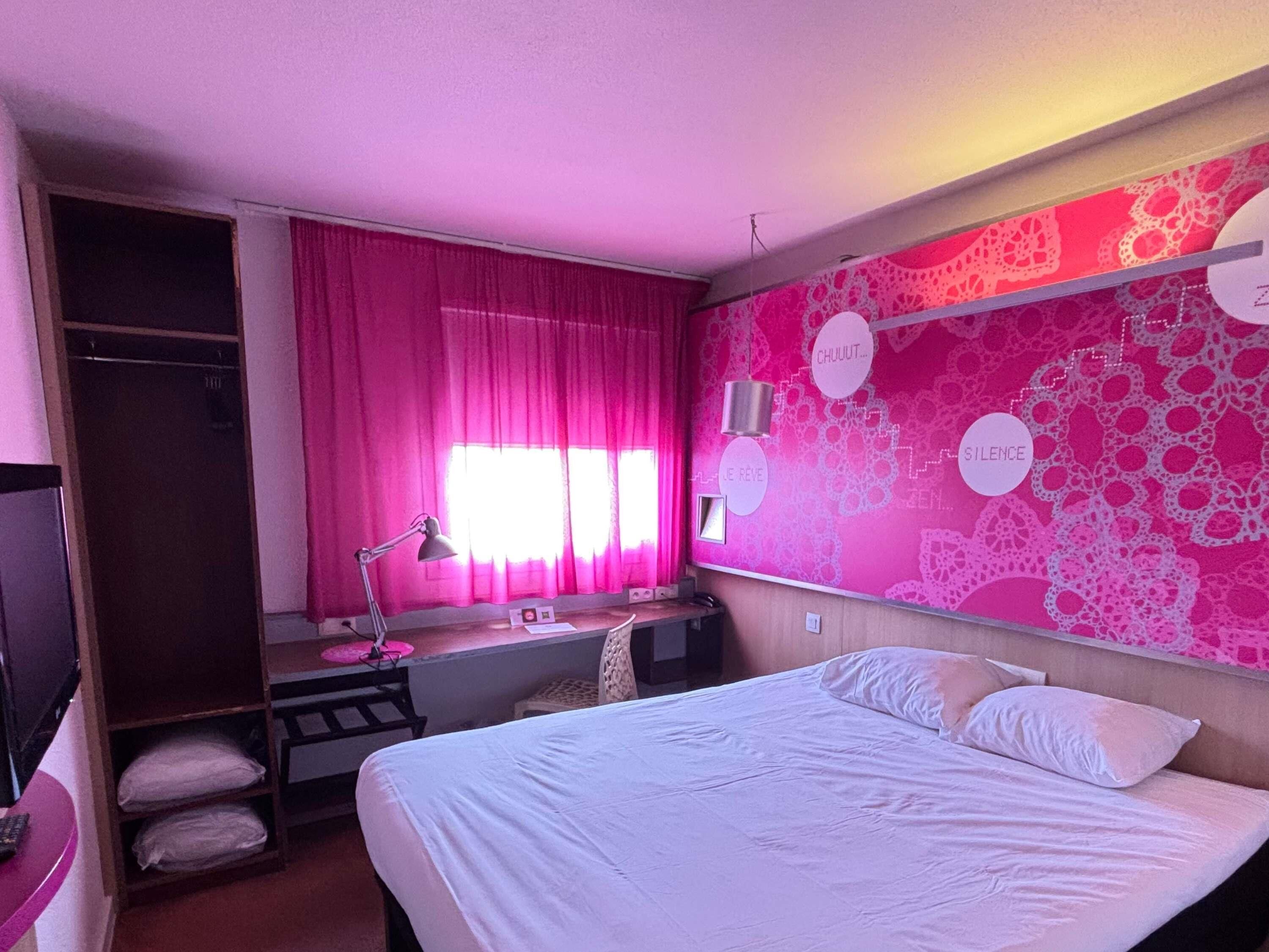 A hotel room with a bed, a desk with a lamp, a wardrobe, and a window with curtains.