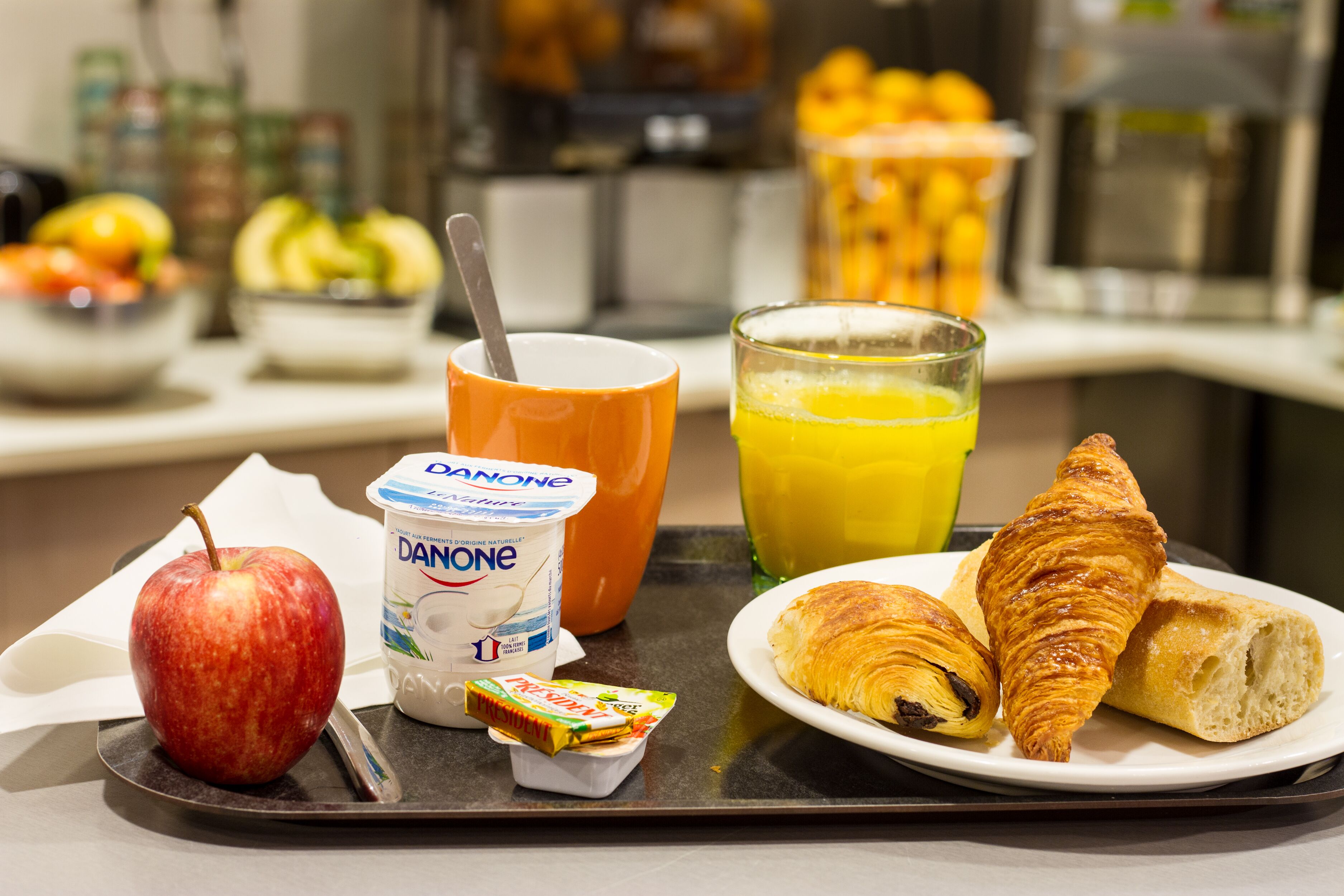 Una bandeja de desayuno con una manzana, yogur Danone, un croissant y un vaso de jugo de naranja.