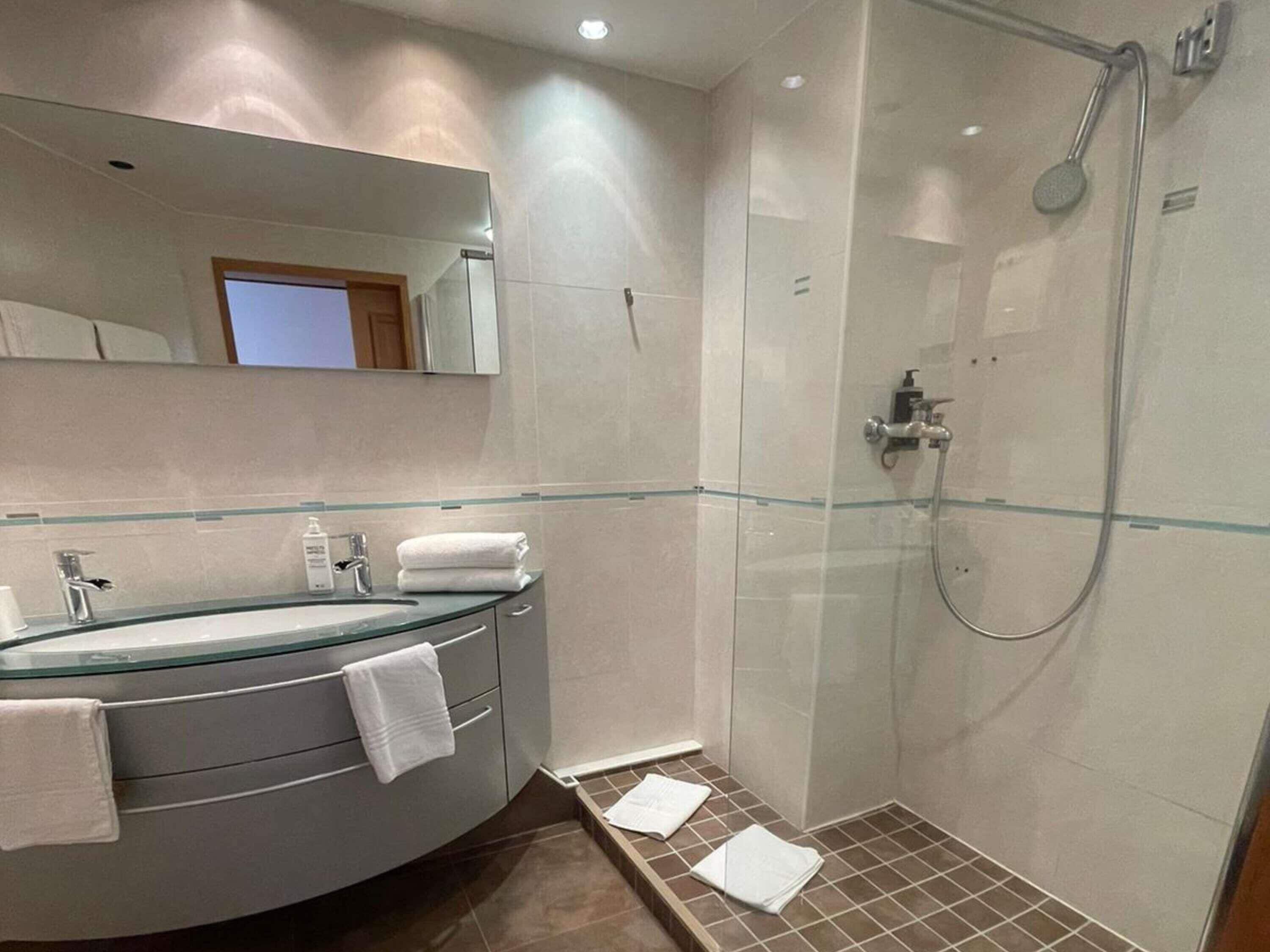 Un baño moderno con ducha de vidrio, tocador curvo con lavabo de vidrio y un espejo grande sobre el lavamanos.