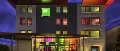 A modern hotel building with a prominent 'ibis Styles Hotels' sign, large windows, and a covered entrance area.
