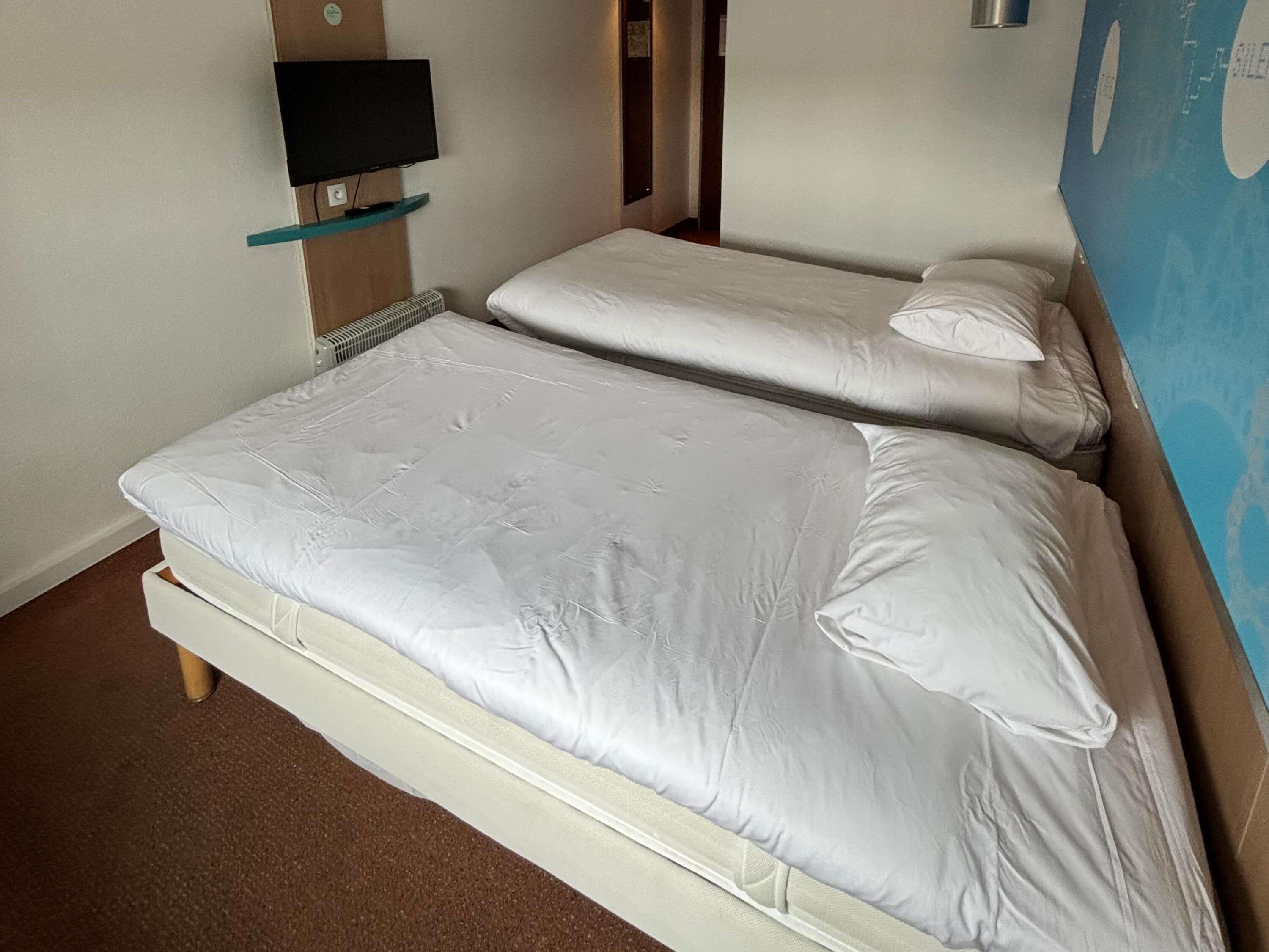A double bed with white bedding, a television mounted on the wall, and a blue wall with a geometric pattern.