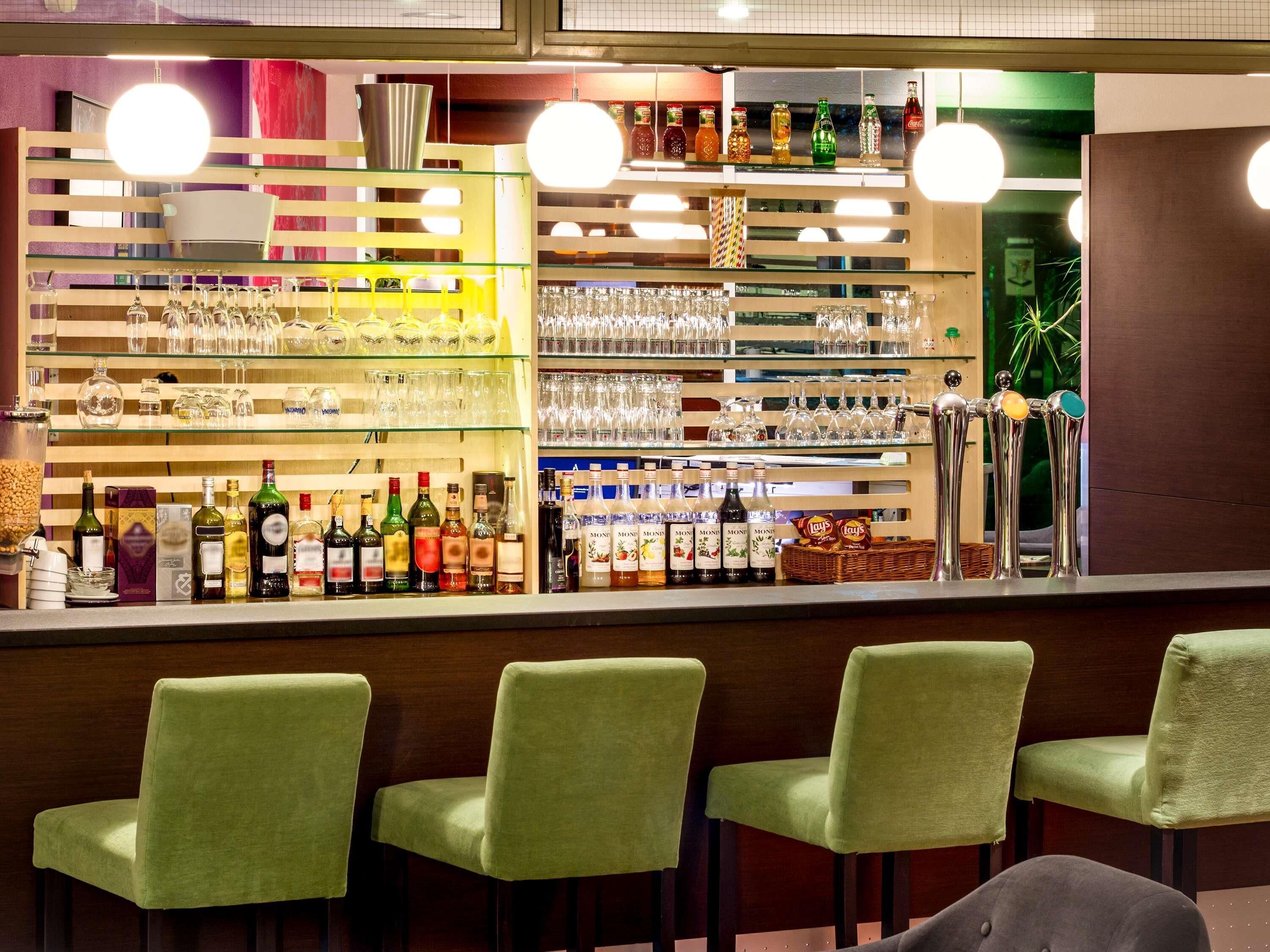 A bar area with glass shelves displaying bottles and glasses, and green upholstered bar stools.