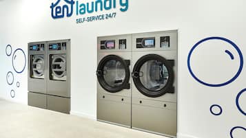 Una lavandería autoservicio con dos lavadoras y secadoras, un panel de control y un cartel en la pared que indica disponibilidad las 24 horas del día, los 7 días de la semana.
