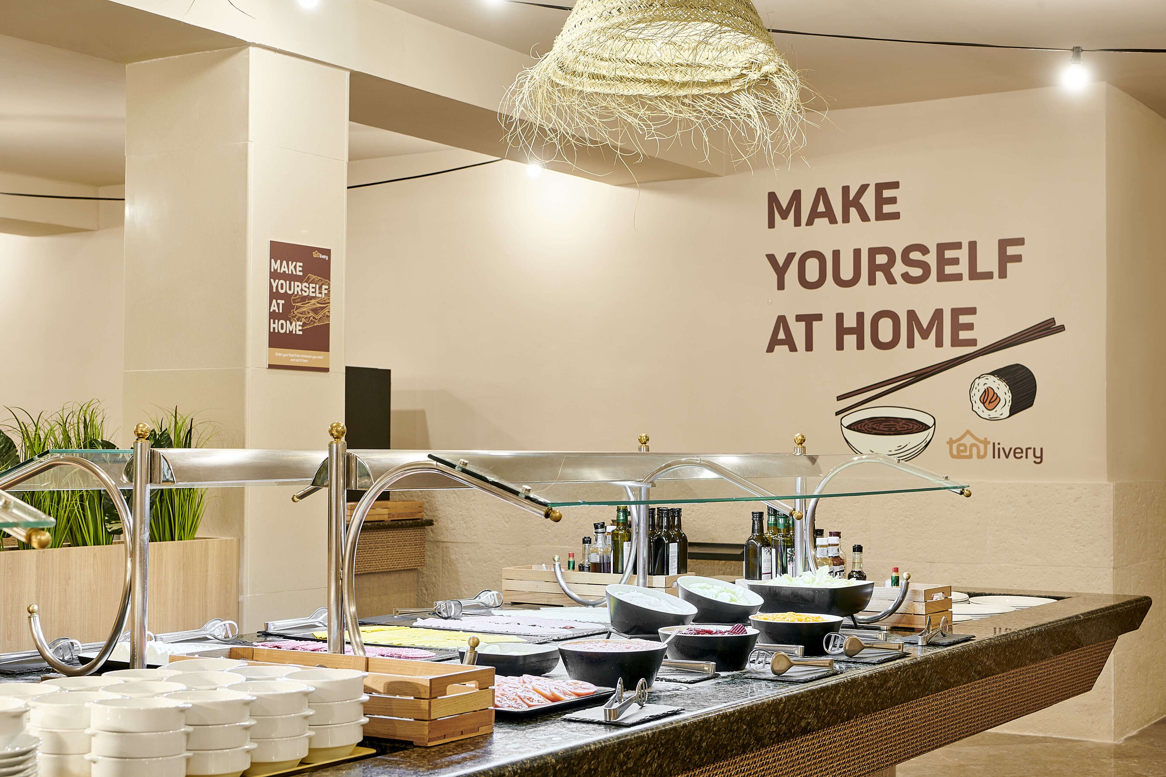 Un montaje de buffet con varios recipientes de comida y utensilios, un letrero que dice 'Hazte con sentir como en casa' y el logotipo de 'tenlivery'.