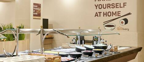 A buffet setup with various food containers and utensils, a sign saying "Make Yourself at Home", and a logo for 'tenlivery'.
