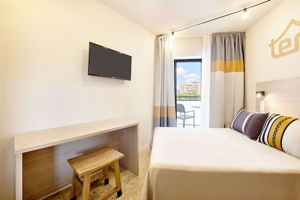 Habitación de hotel con cama, escritorio, televisor y balcón con cortinas.