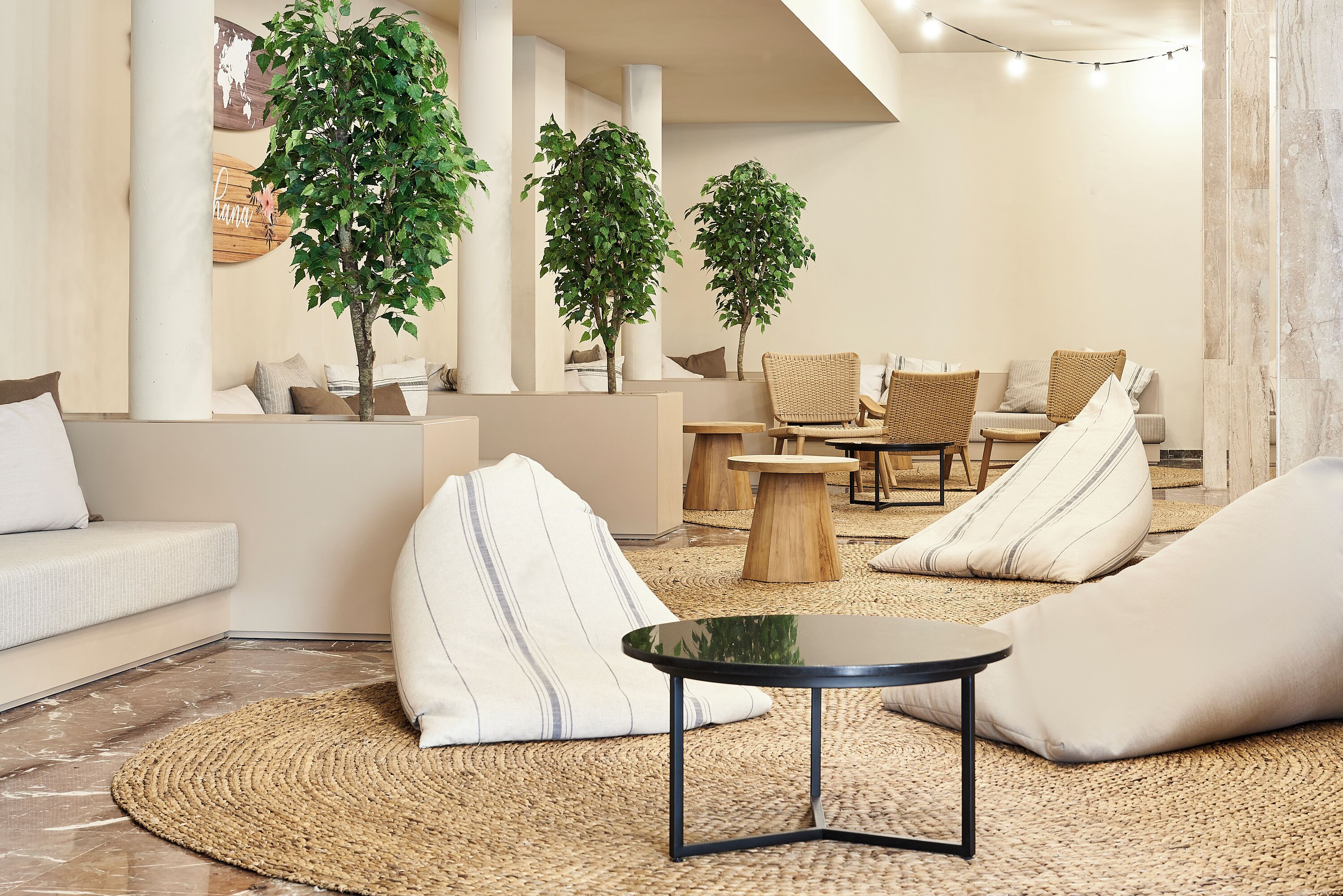 A modern lounge area with a sofa, bean bags, a round table, and potted plants.