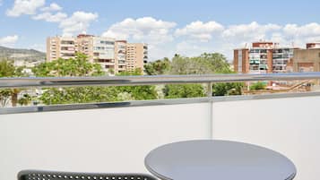 A balcony with a round table and a chair, overlooking a cityscape with multi-story buildings and greenery.