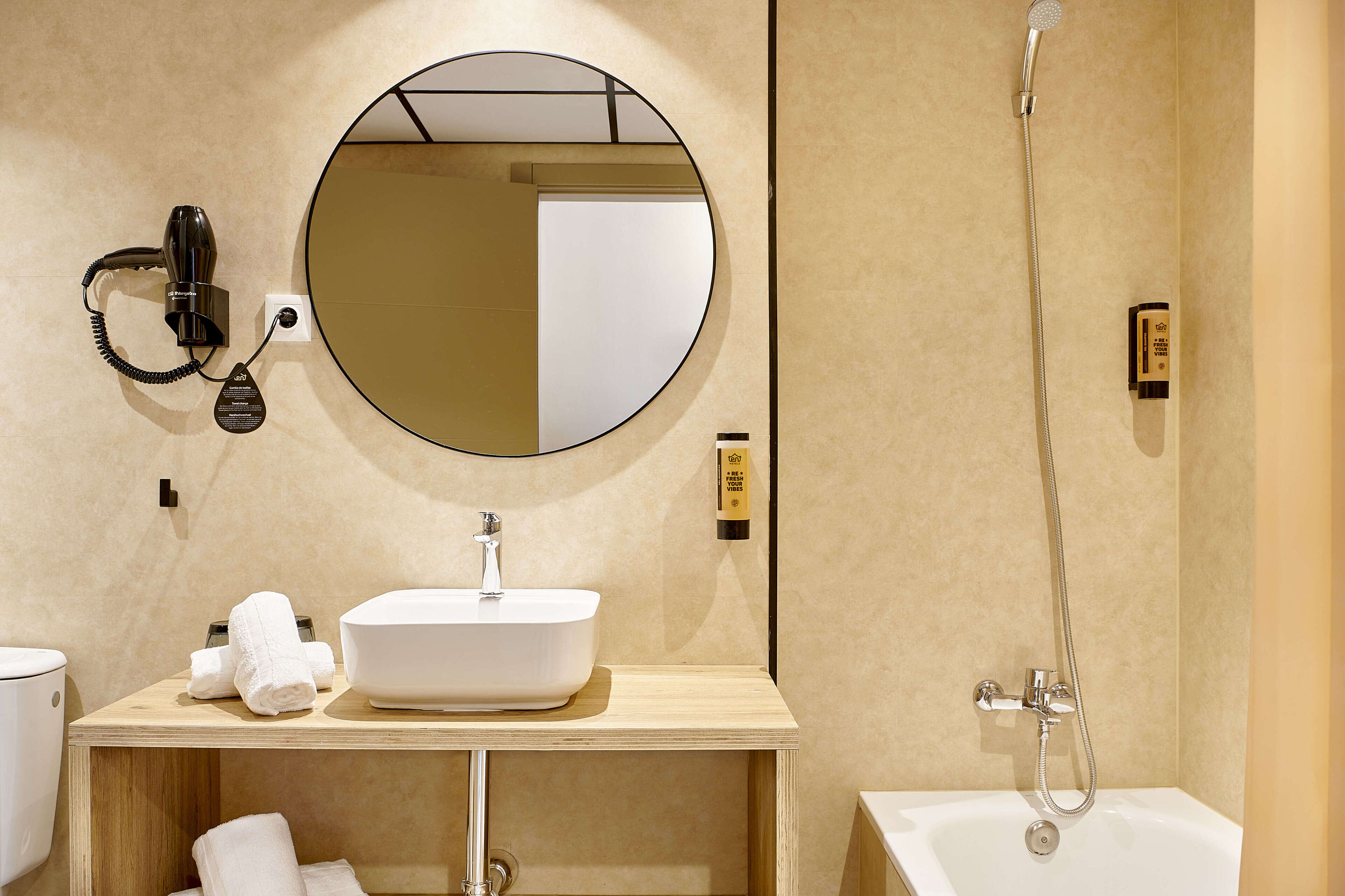 Un bagno moderno con uno specchio rotondo, un lavabo bianco, un asciugacapelli portatile, un soffione doccia manuale e una vasca da bagno.