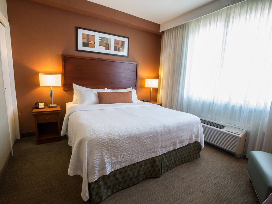 A hotel room with a large bed, two bedside tables with lamps, a headboard with a framed picture, a window with curtains, and a carpeted floor.