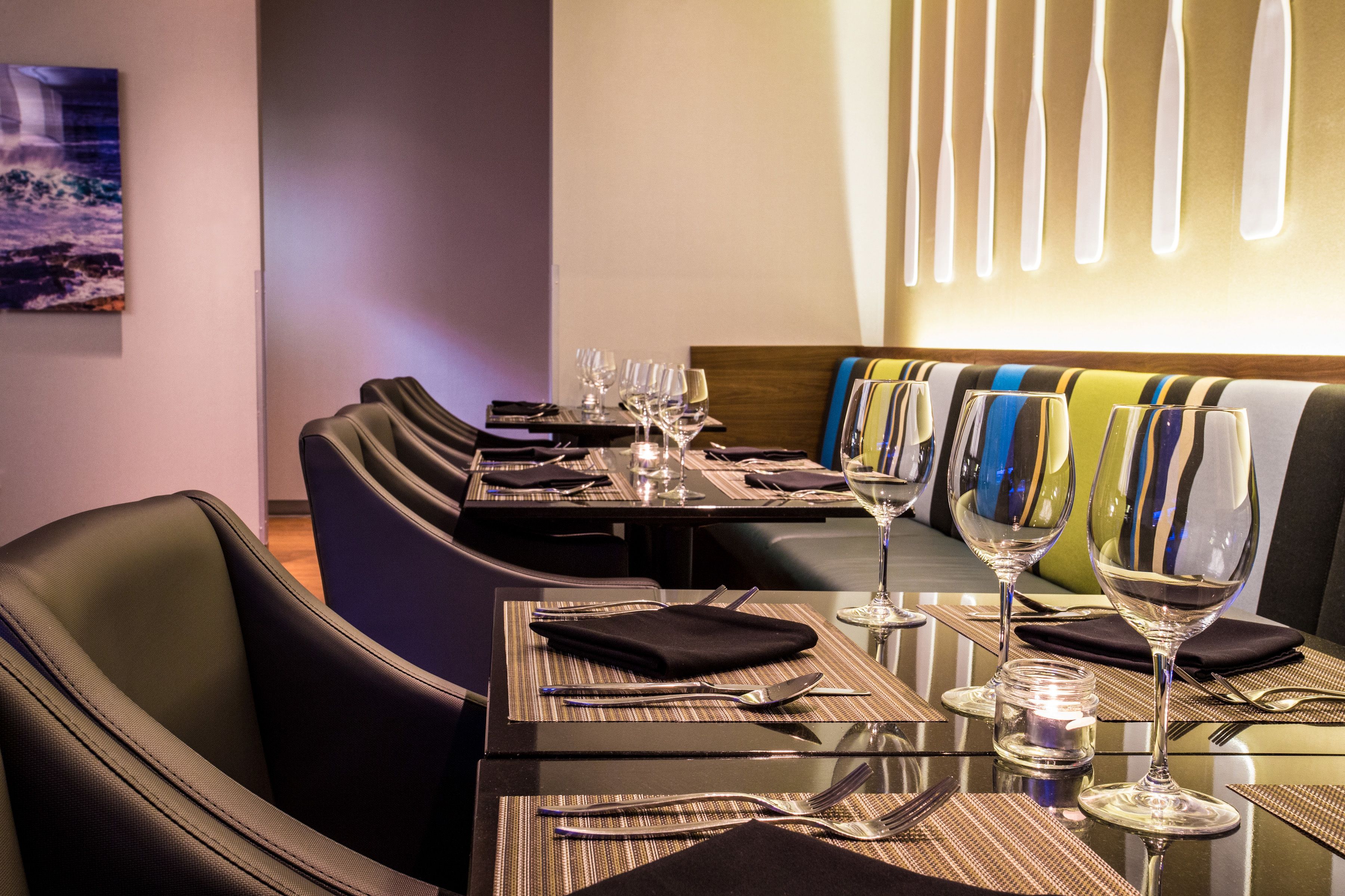 Un comedor con mesas dispuestas para cuatro, cada una con un mantel individual, cubiertos y copas de vino. Hay sillas acolchadas y un sofá con cojines a rayas.