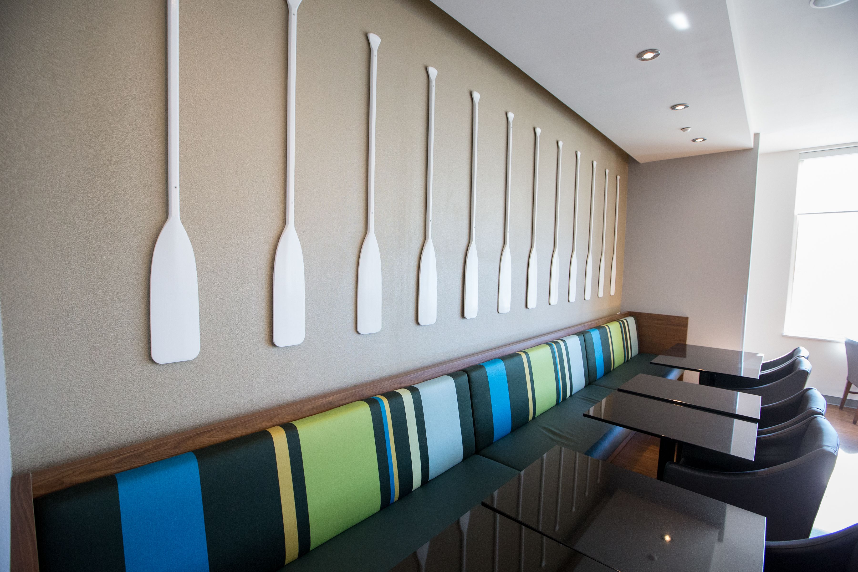 A modern interior with a long bench with striped cushions, a wall-mounted row of white paddles, and a glass-top table with chairs.