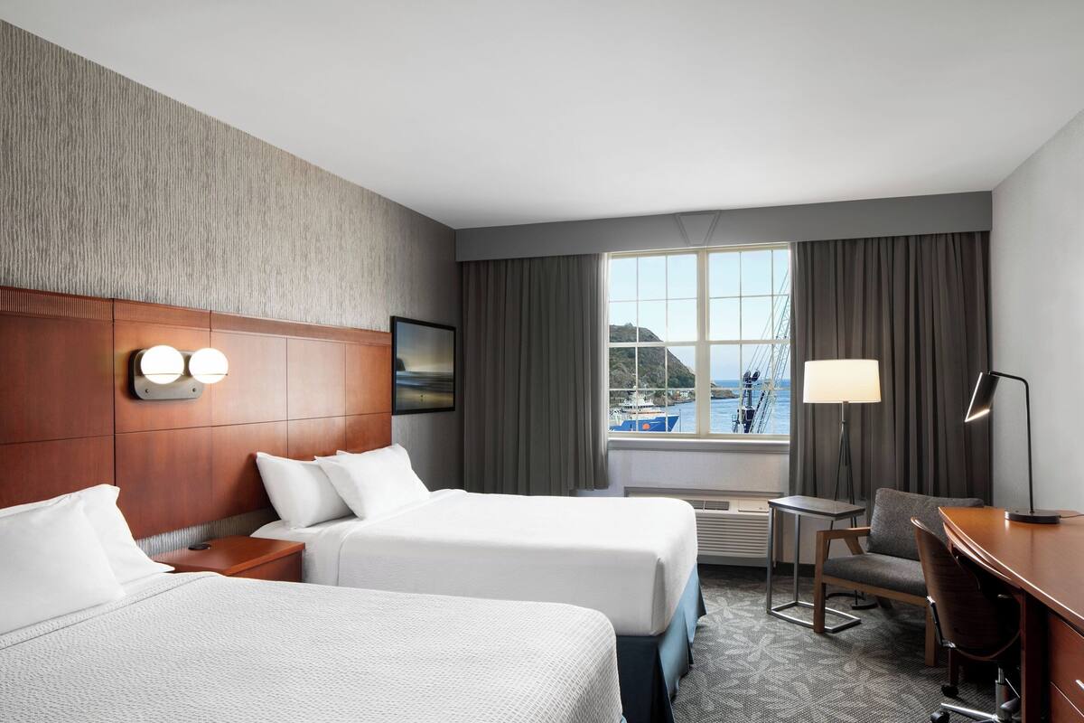 A hotel room with two beds, a desk, a chair, a lamp, a view of the sea, and a window with curtains.