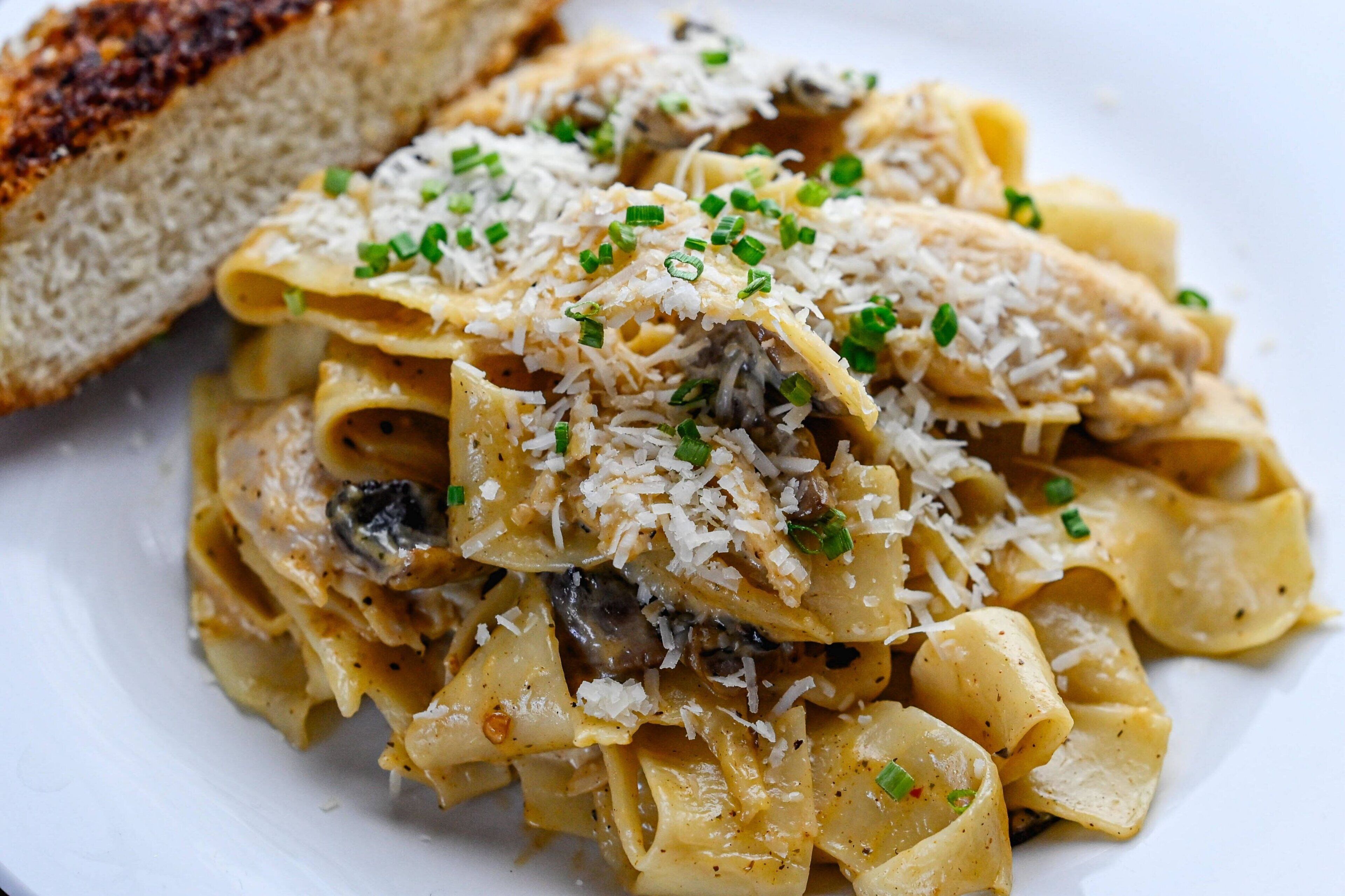 Une assiette de pâtes aux champignons, recouverte de fromage râpé, accompagnée d’une tranche de pain grillé.