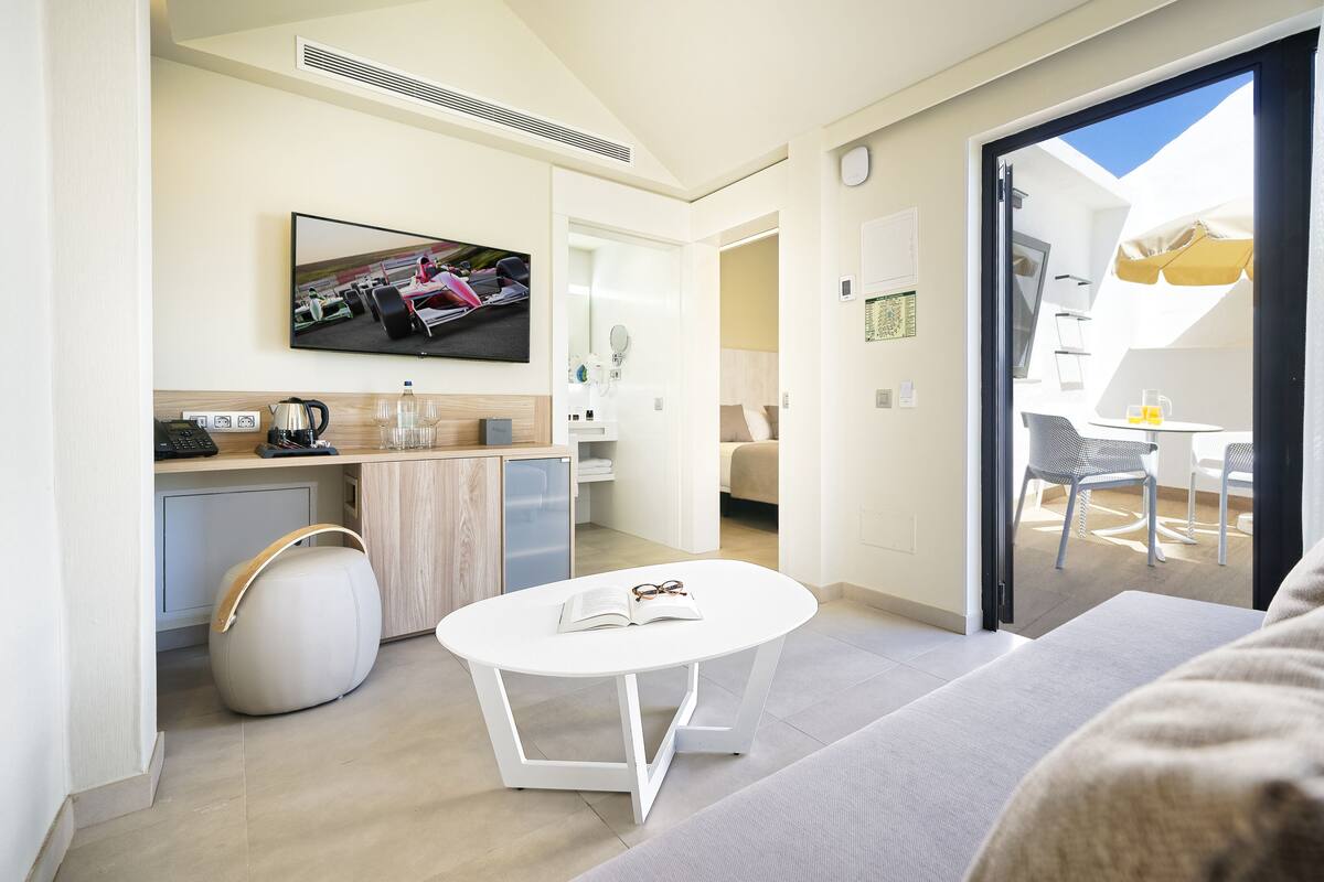 Ein modernes Hotelzimmer mit einer Couch, einem kleinen Esstisch, einer Küchenzeile, einem Fernseher und einem Balkon mit Tisch und Stühlen.