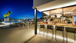 A bar area with a poolside seating arrangement, a bar counter with various bottles, and a view of the pool at dusk. - Club Maspalomas Suites & Spa - Adults Only (San Bartolomé de Tirajana)