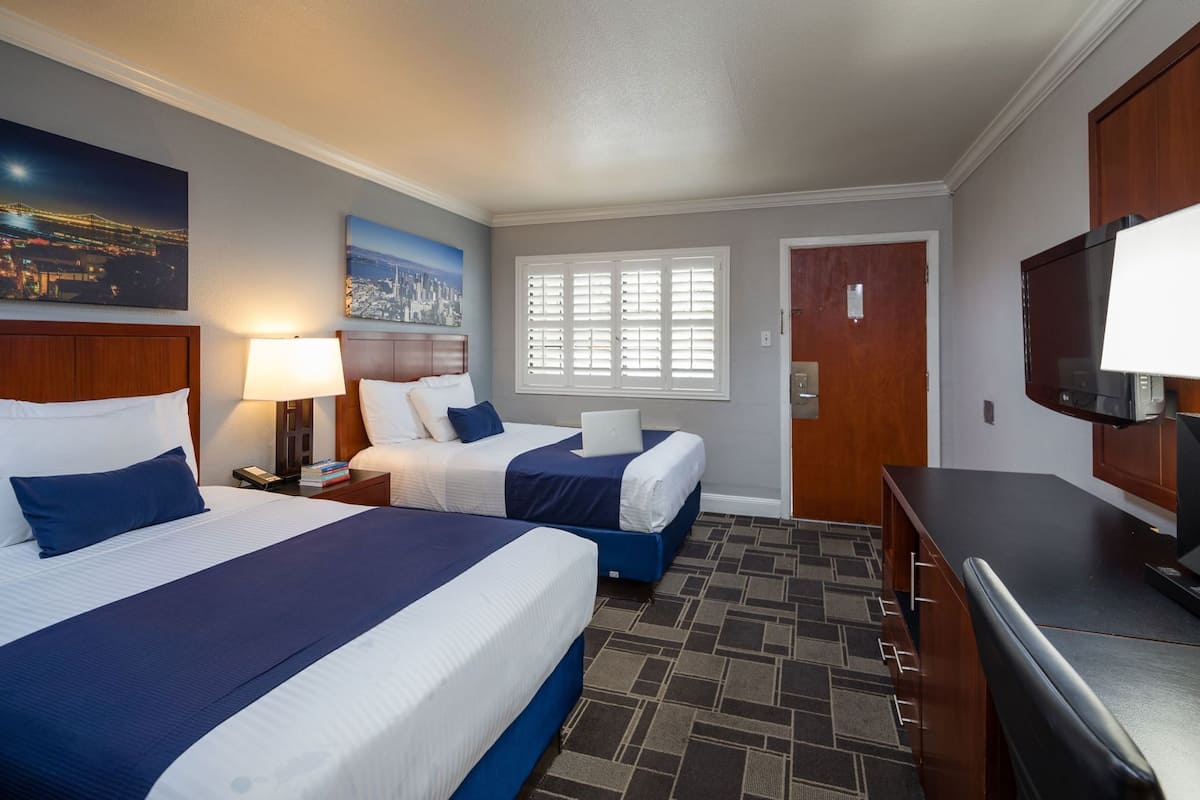 A hotel room with two beds, a desk, a TV, a window with blinds, and a painting on the wall.