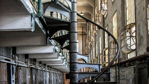 Un escalier en colimaçon dans un bâtiment de style industriel, à la lumière tamisée, avec des rampes en métal et des fenêtres grillagées.