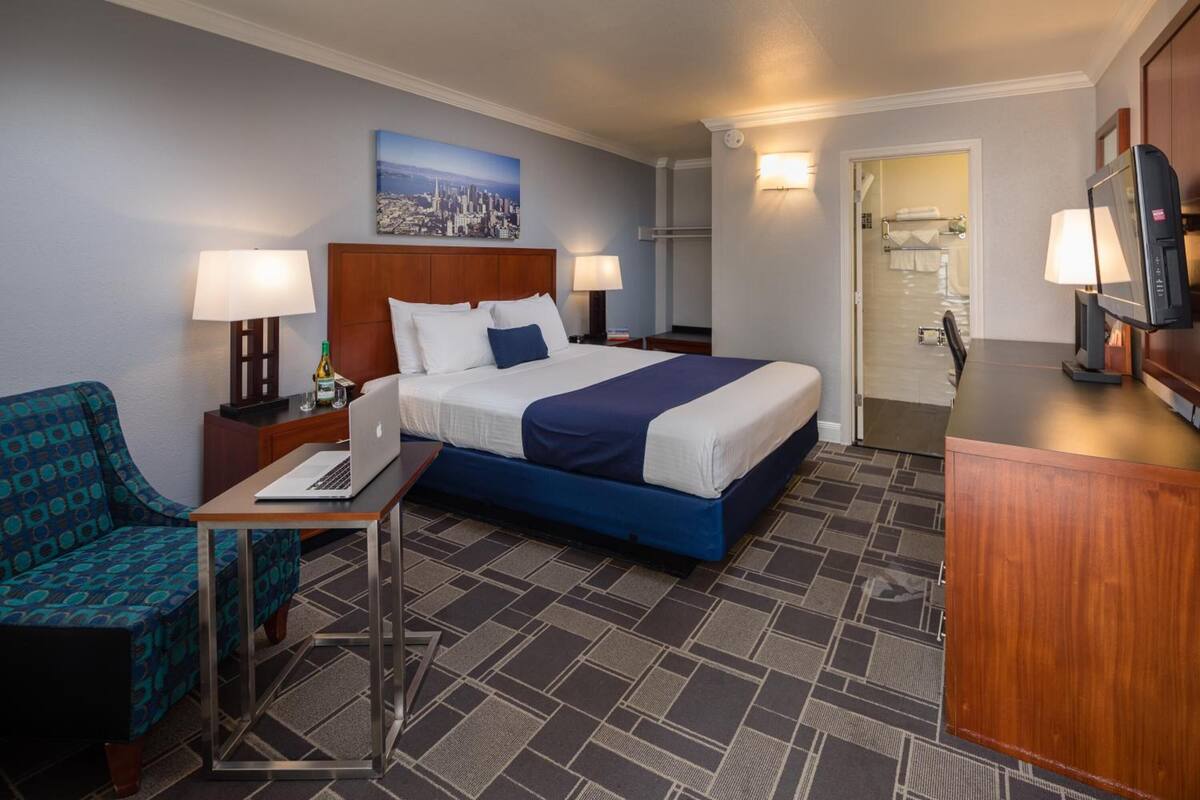 A hotel room with a bed, a desk, a chair, a TV, and a bathroom visible through an open door.