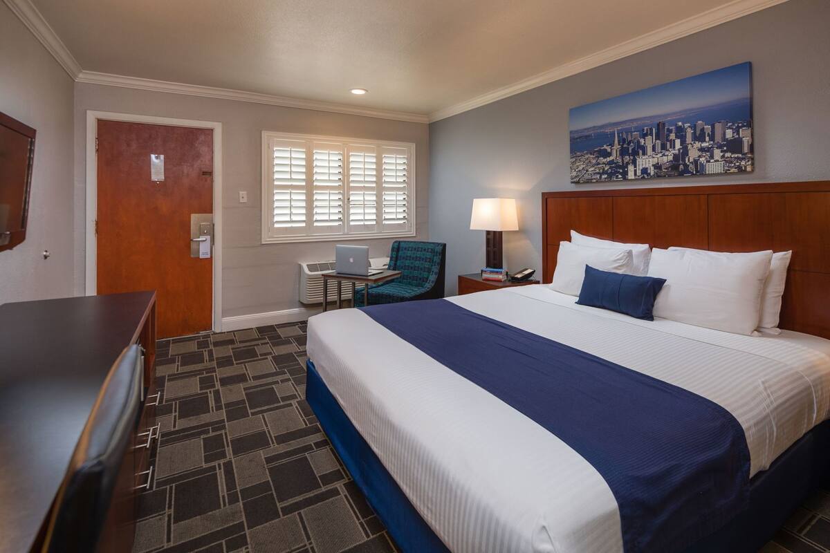 A hotel room with a bed, desk, chair, and a cityscape painting on the wall.