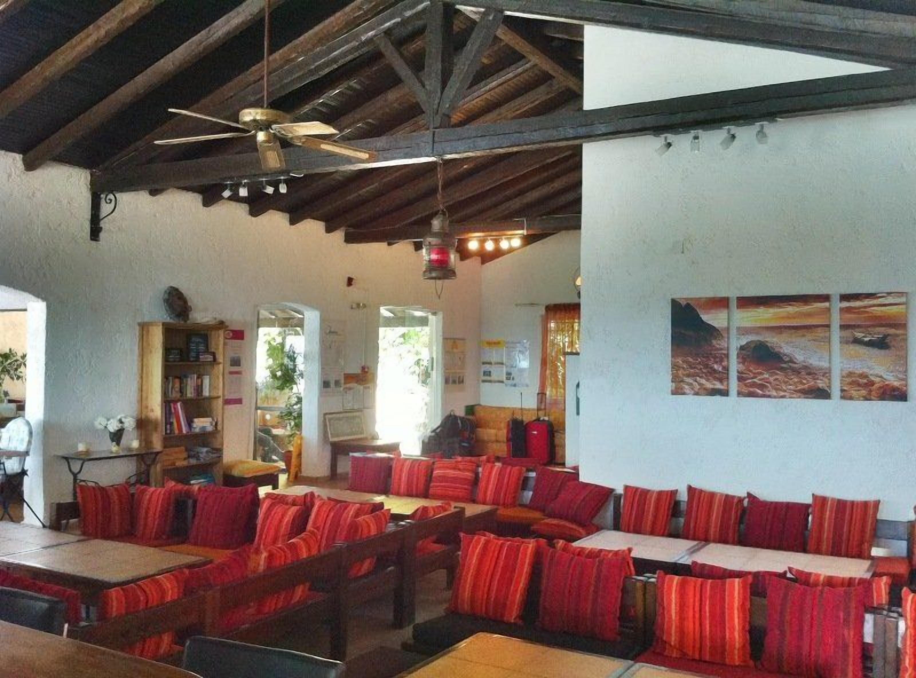 Un restaurante con mesas de madera y cojines a rayas rojas, un ventilador de techo y cuadros en la pared.
