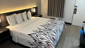 A hotel room with a bed, nightstands, a desk, a chair, a window with curtains, and a door. - Red Roof Inn Wildwood, FL - The Villages (Wildwood)