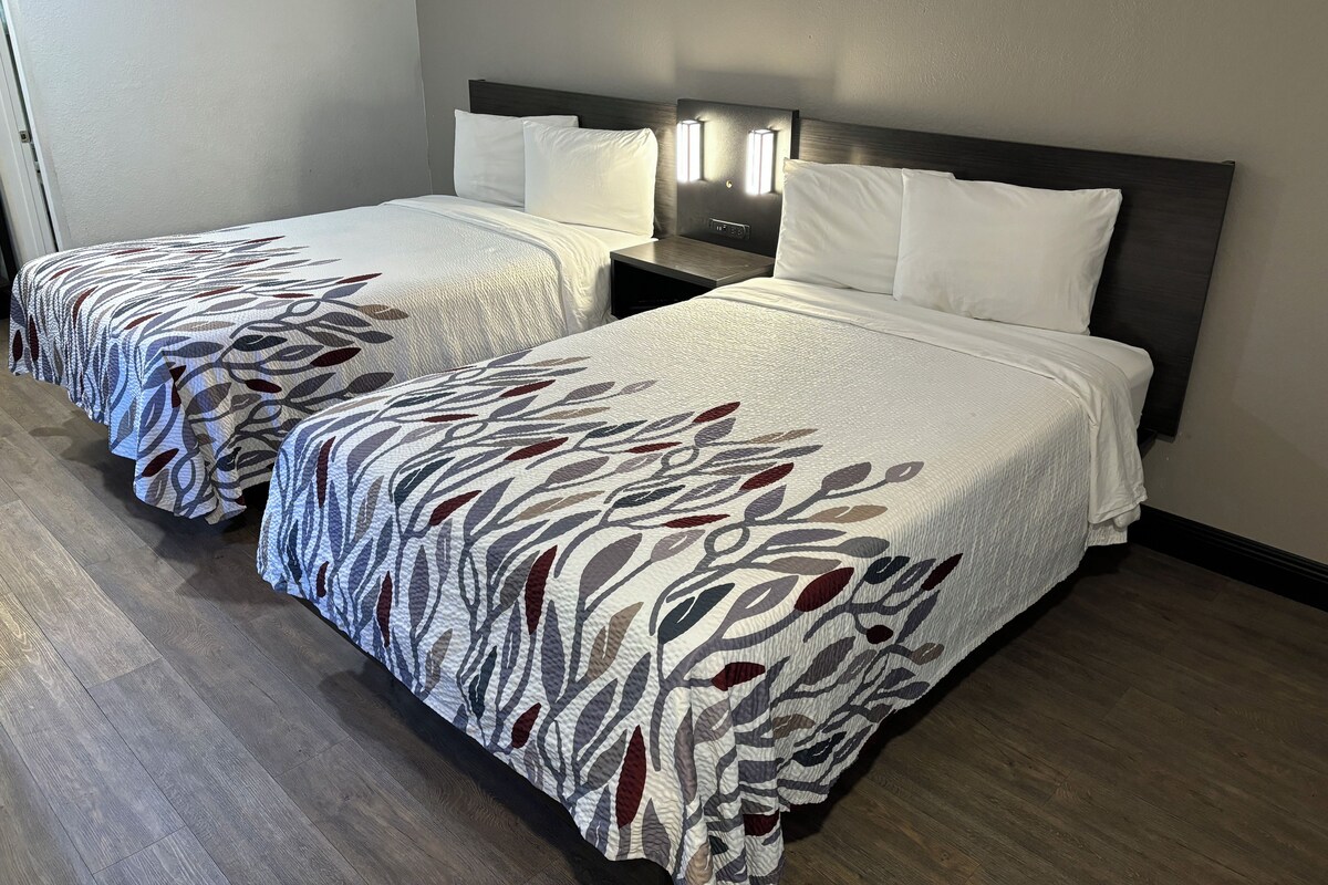 A hotel room with two beds, each with a patterned bedspread, a nightstand with a lamp, and a wooden floor.