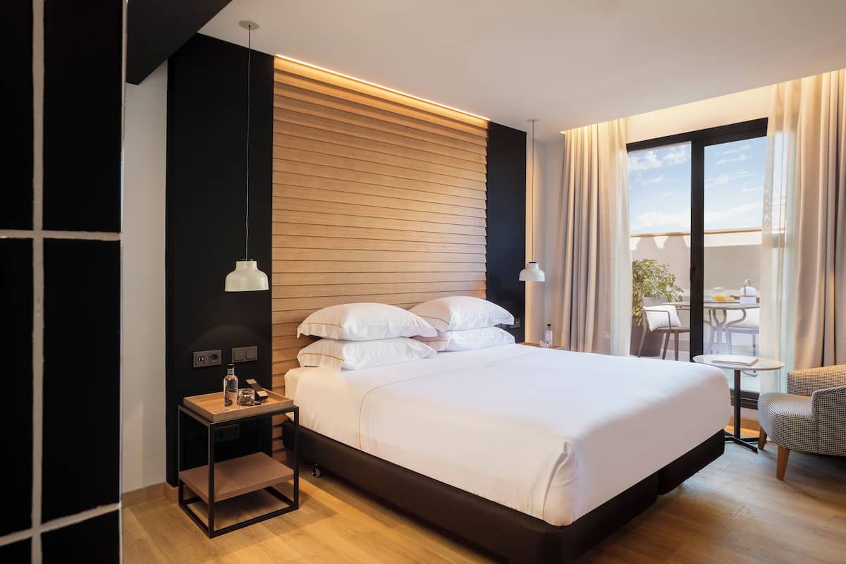 A modern hotel room with a large bed, a small bedside table, a nightstand, a view of the outdoor seating area through a glass door, and a wooden panel wall.