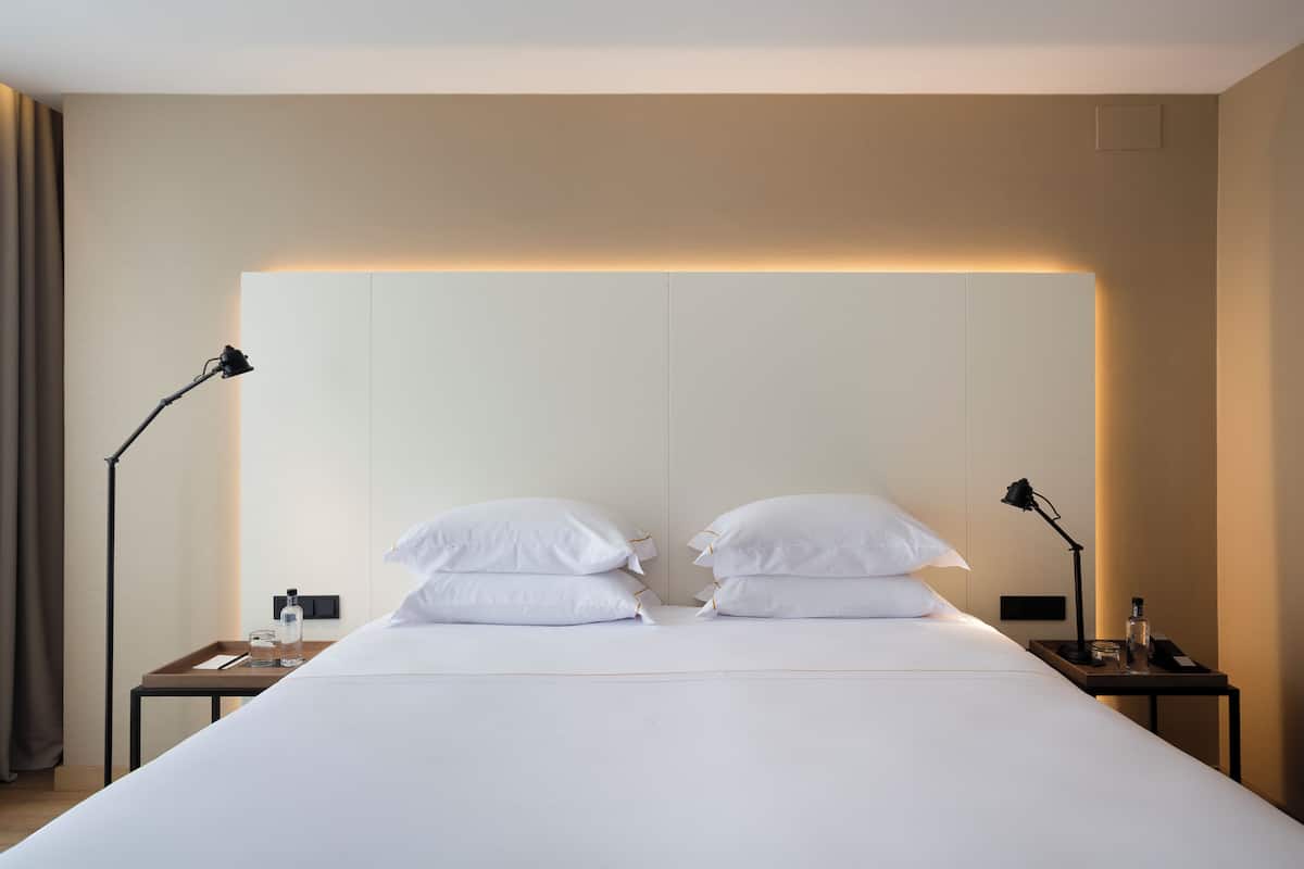 A neatly made bed with white linens and pillows, two bedside tables with lamps, a bottle, and a glass on one of the tables.