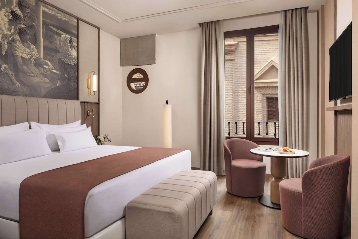 A hotel room with a large bed, a small round table, two chairs, a TV, and a window with curtains.