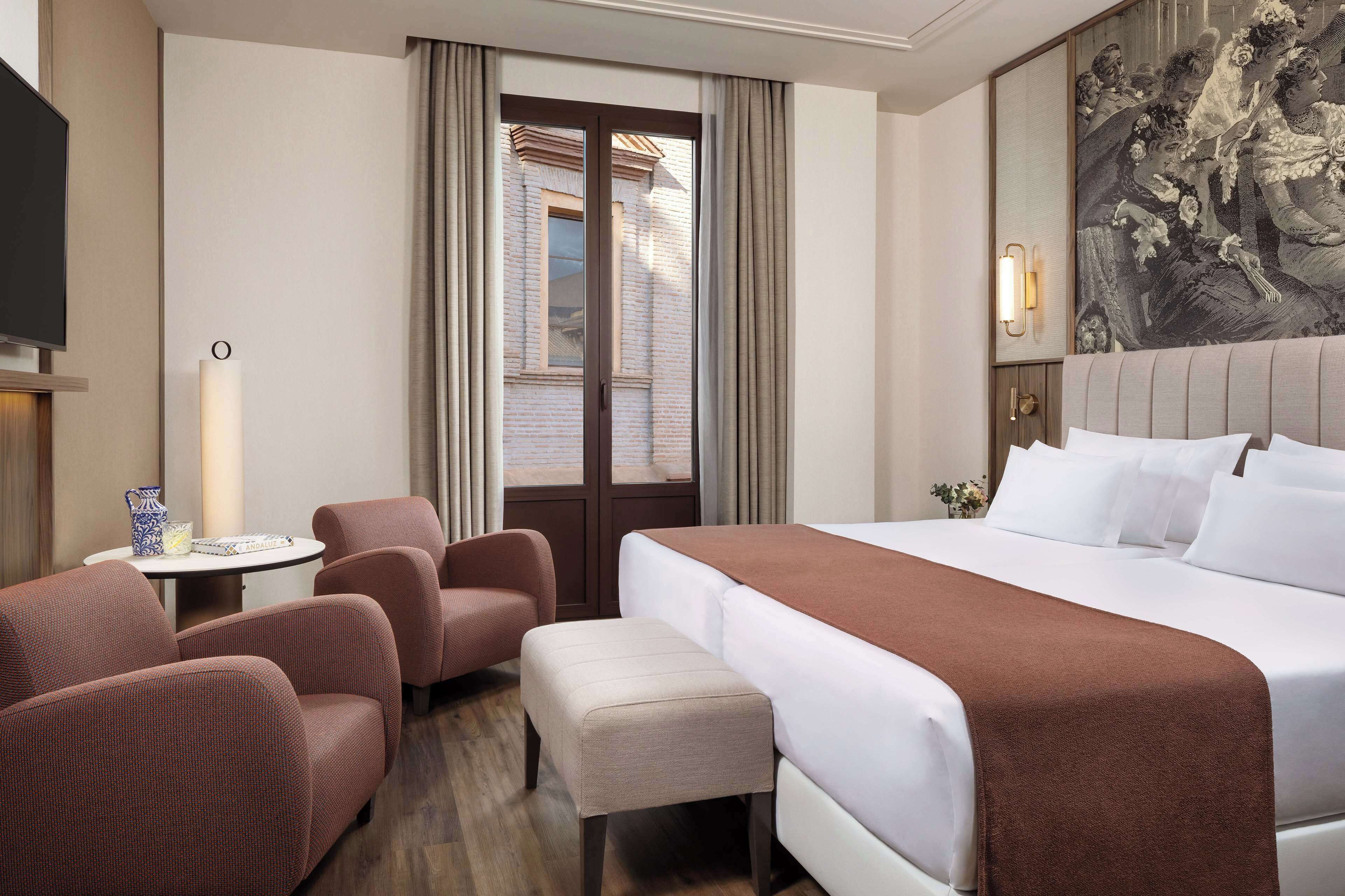 Quarto de hotel com uma cama grande, duas poltronas, uma mesa pequena com um vaso e um quadro na parede.