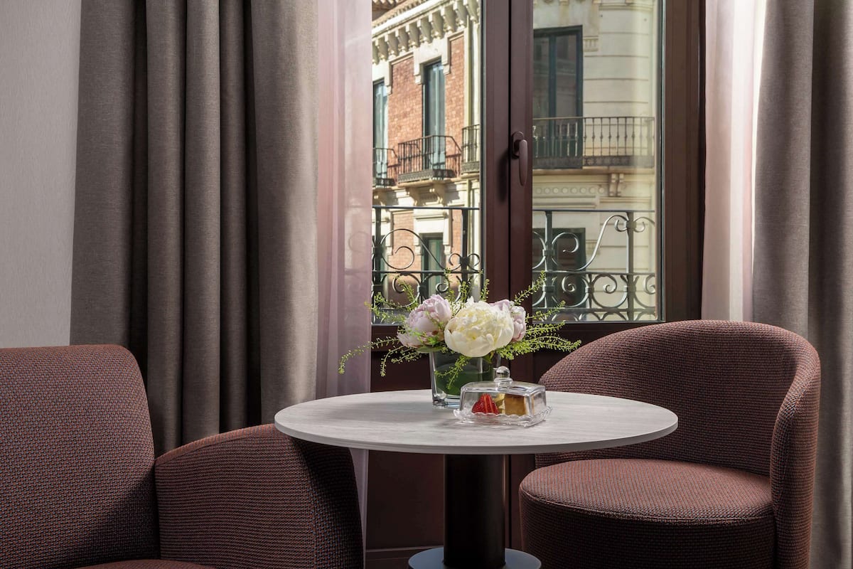 A round table with a vase of flowers and a glass container, set between two upholstered chairs, with a view of a balcony and building through a window.