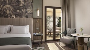 A hotel room with a bed, a small table, a chair, a large window, and a mural on the wall.
