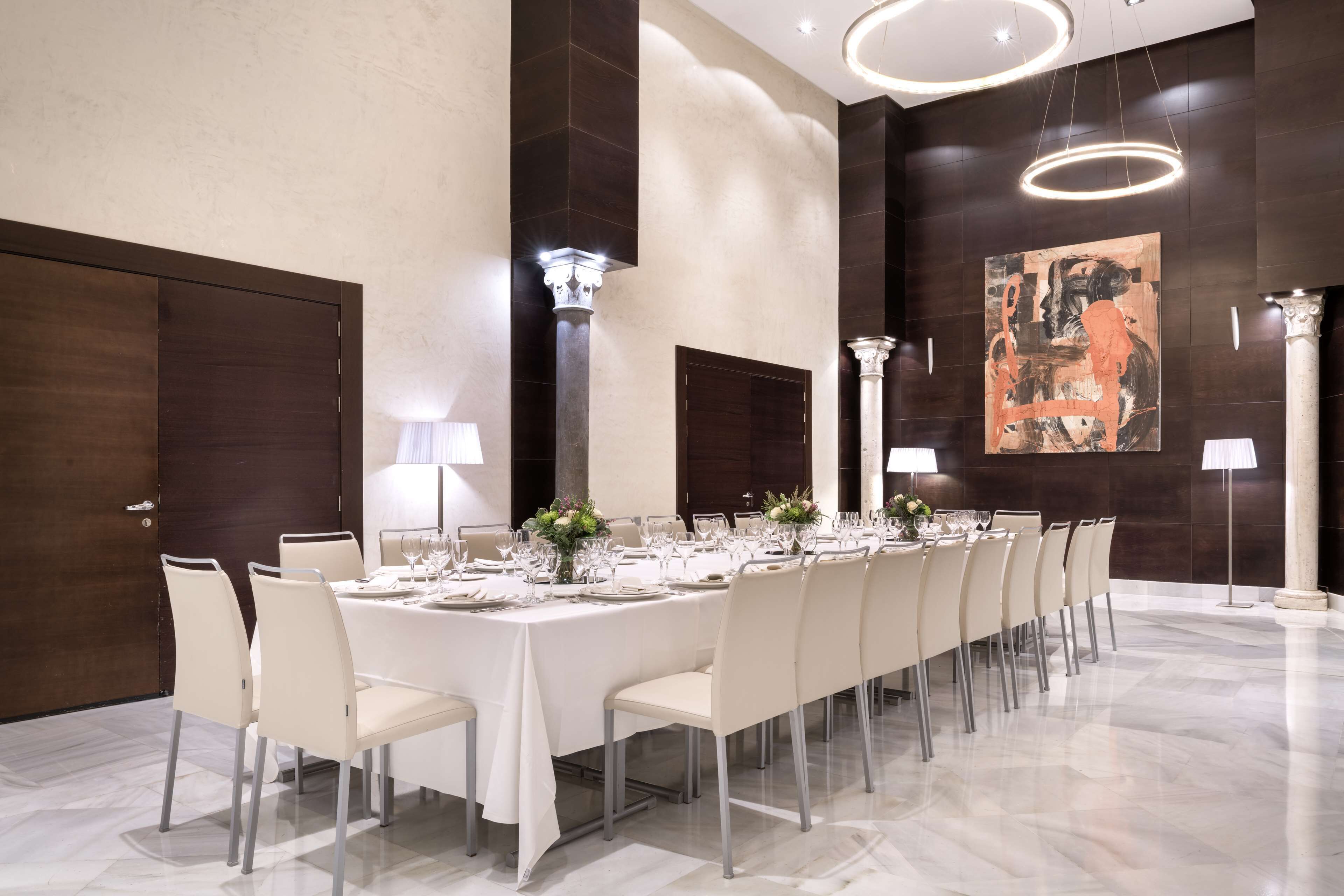 Una larga mesa de comedor dispuesta para una comida formal, rodeada de sillas, con una lámpara moderna y obras de arte en las paredes.