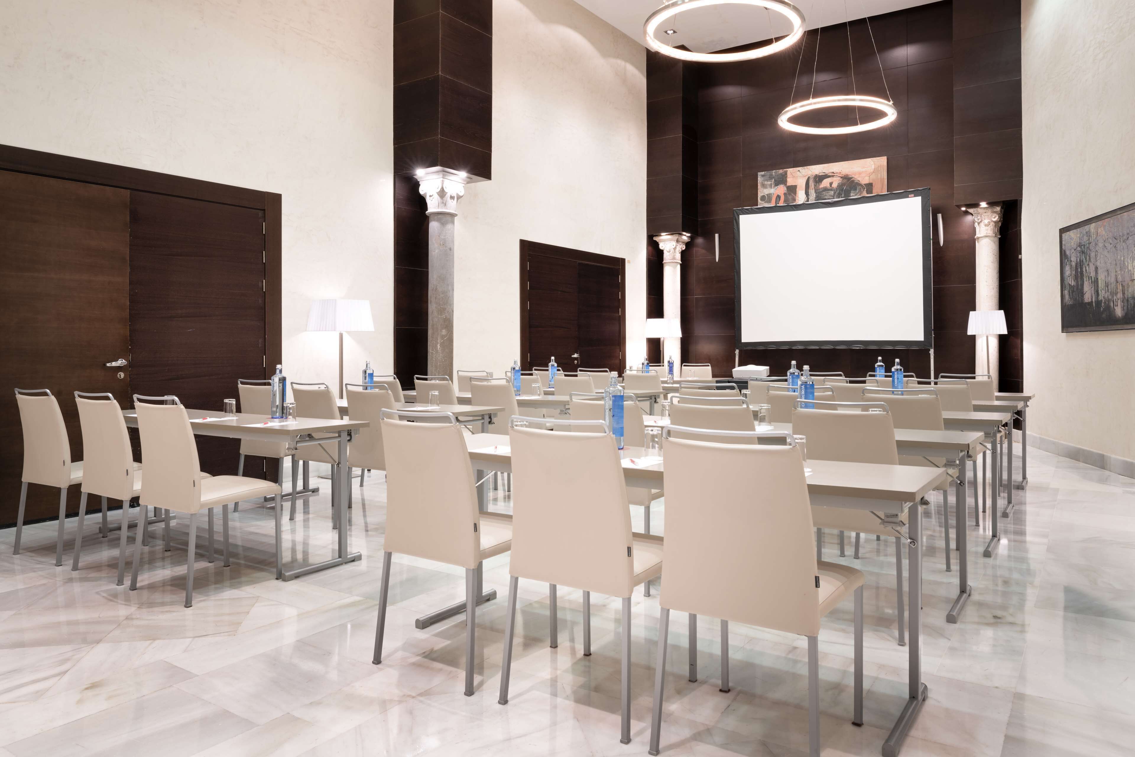 A conference room with tables and chairs, a projector screen, and a large screen on the wall.