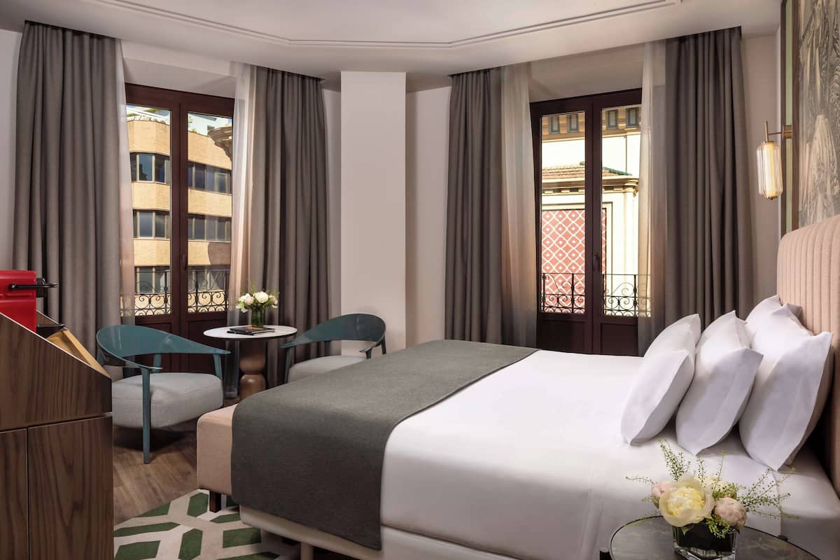 A hotel room with a bed, a desk, a chair, and a view of buildings.