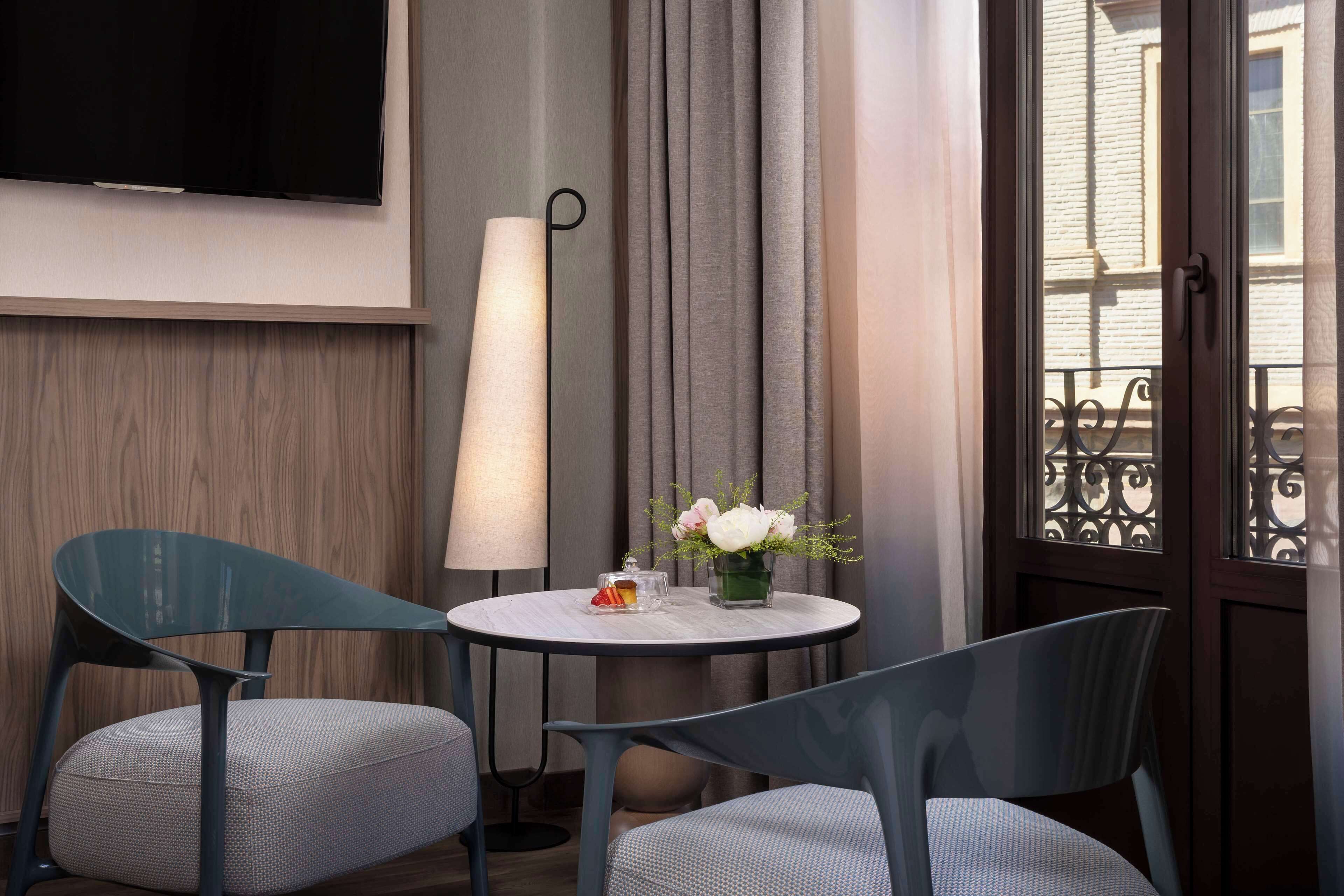 Habitación de hotel con una mesa redonda, dos sillas, un televisor de pantalla plana, una lámpara de pie y una ventana con vista a un edificio.