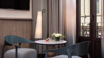 Habitación de hotel con una mesa redonda, dos sillas, un televisor de pantalla plana, una lámpara de pie y una ventana con vista a un edificio.