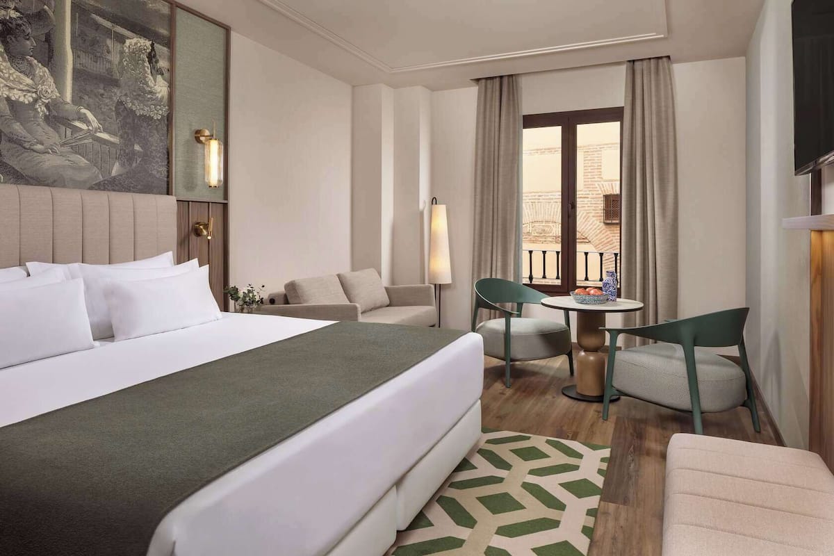 A hotel room with a large bed, a sofa, a dining table with chairs, and a view of the outside.