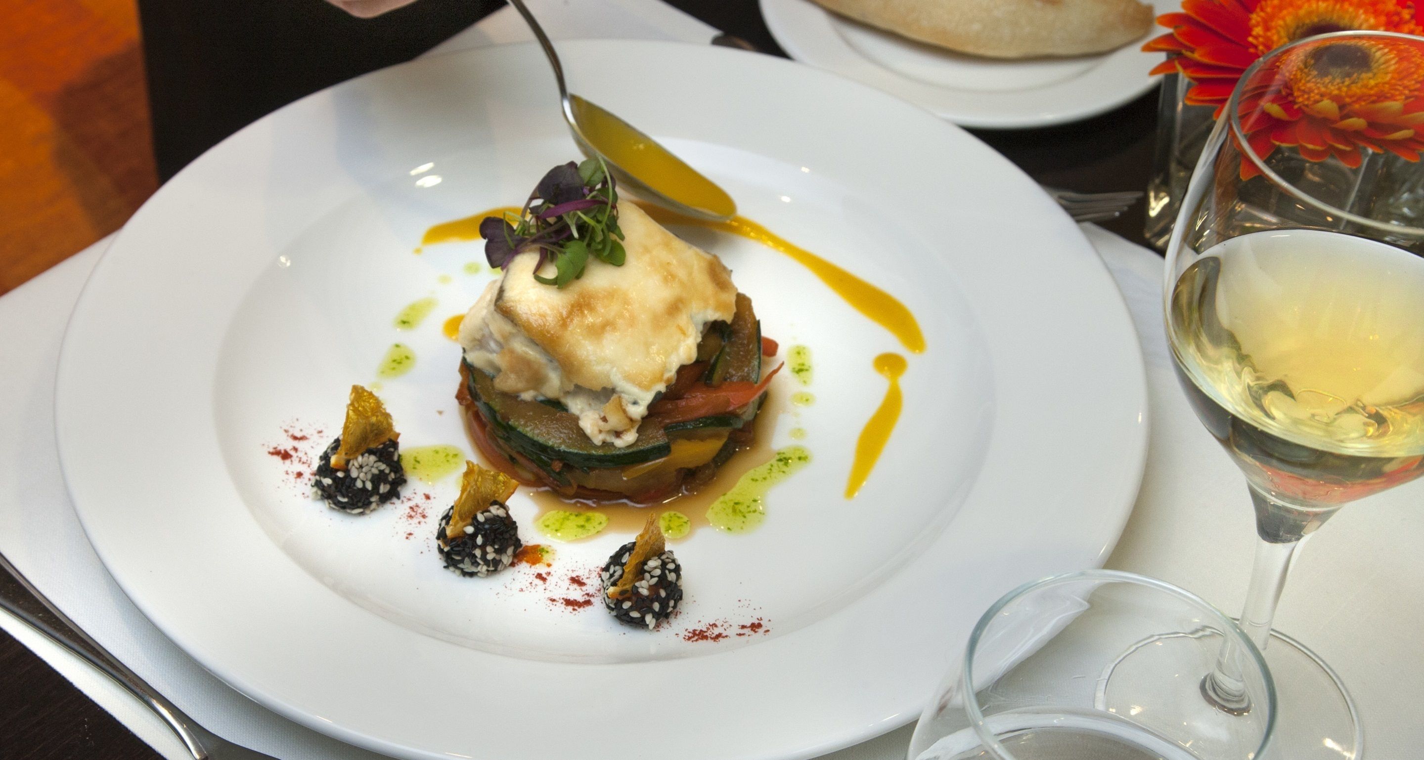 Une assiette blanche contenant un plat gastronomique, un verre de vin blanc et un verre d’eau, le tout posé sur une table.