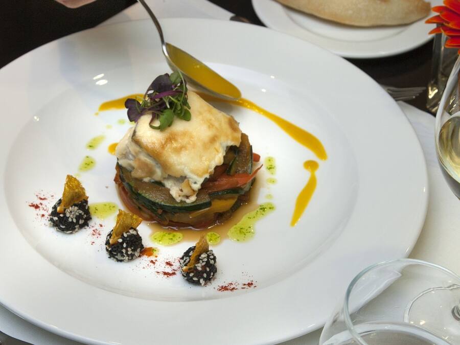 Un plato blanco con un plato gourmet, una copa de vino blanco y un vaso de agua sobre una mesa.