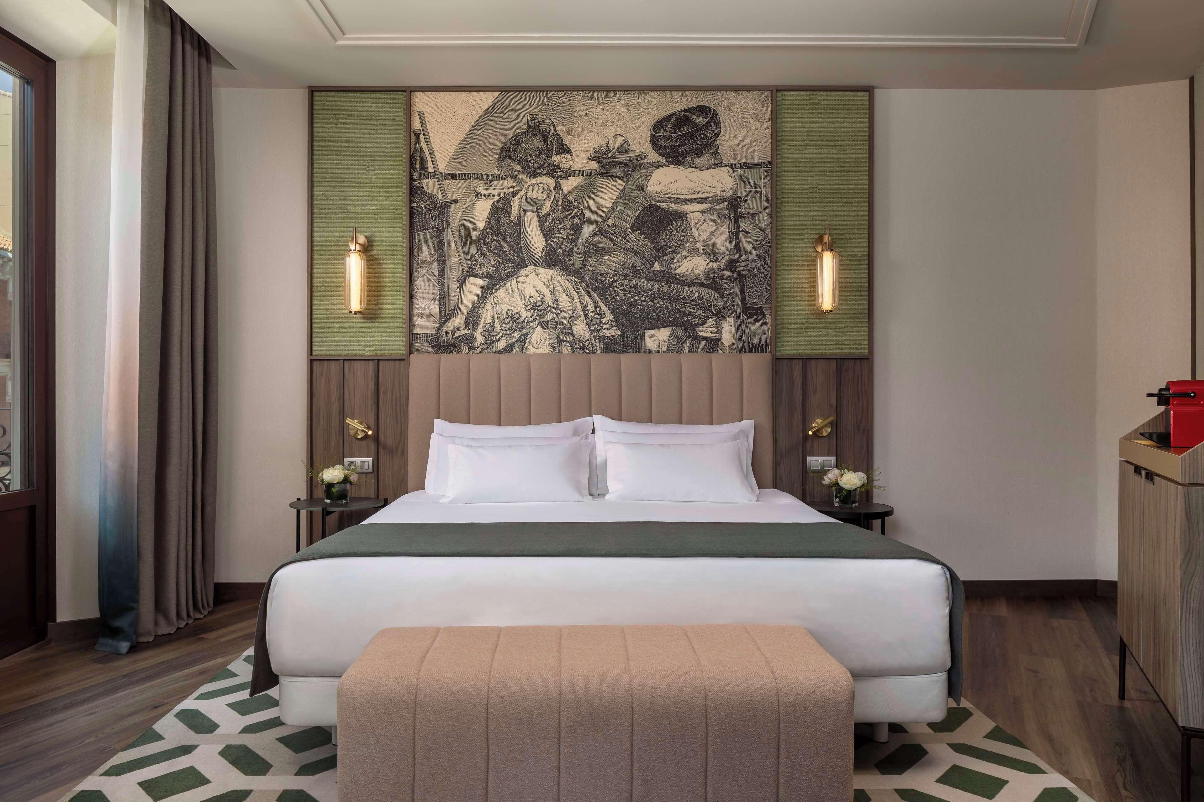 A hotel room with a large bed, a headboard with artwork, two bedside tables with lamps, and a window with curtains.