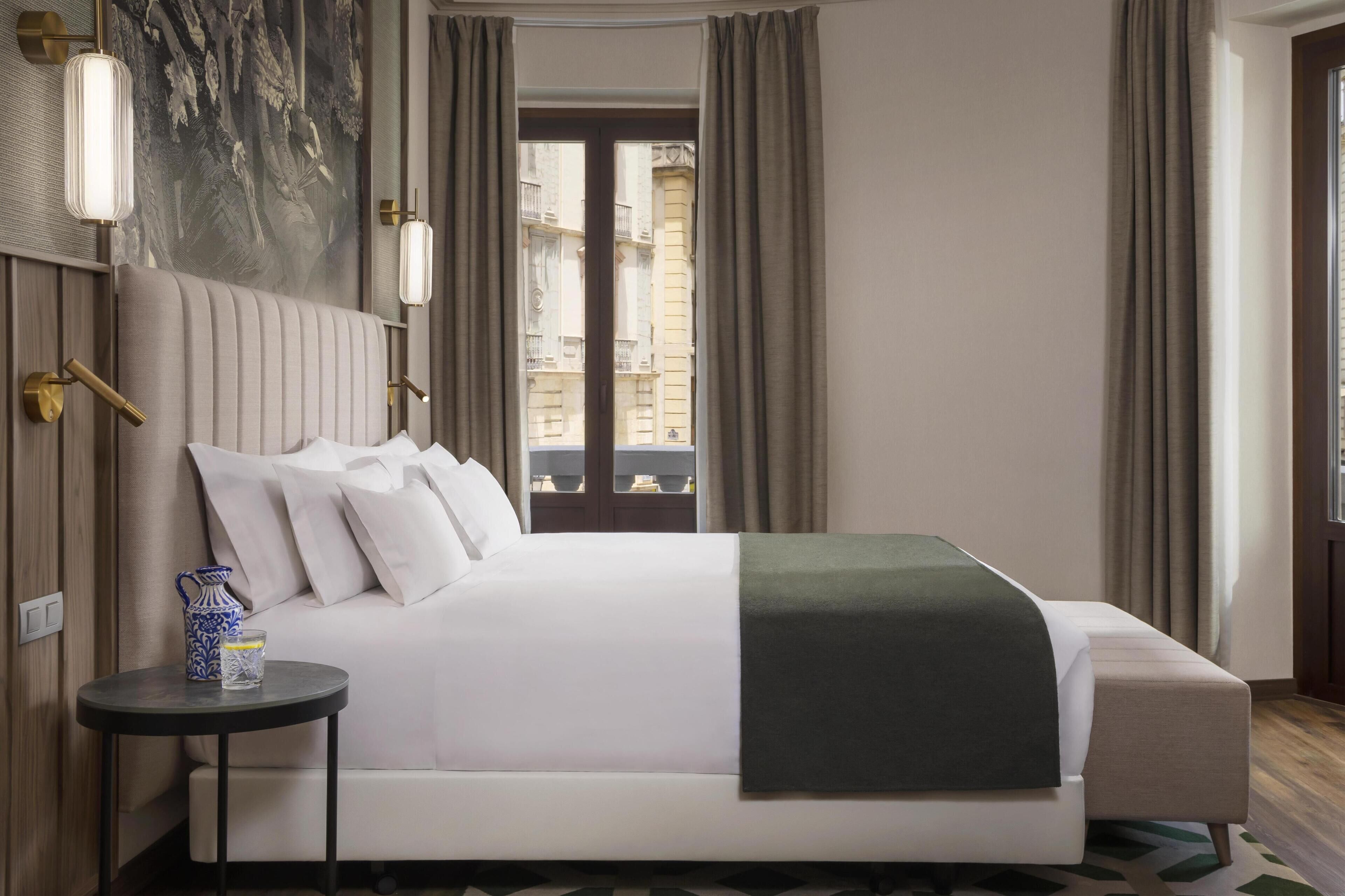 Quarto de hotel com uma cama grande, uma mesinha com um jarro e vista para edifícios pela janela.