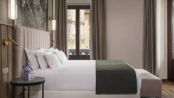 Quarto de hotel com uma cama grande, uma mesinha com um jarro e vista para edifícios pela janela.