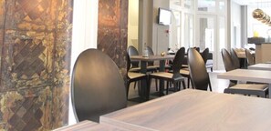 A modern restaurant interior with wooden tables and black chairs, a stone wall, and a television on the wall.