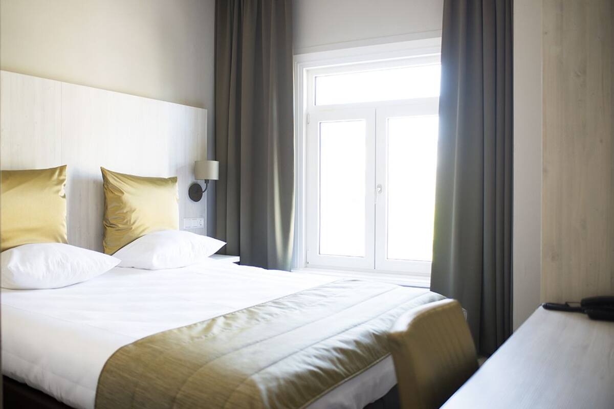 Hotellihuone, jossa on suuri sänky, yöpöytä, lamppu ja ikkuna, jossa on verhot.