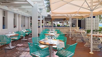 Ein Restaurant mit Außenbereich, ausgestattet mit türkisfarbenen Stühlen und weißen Tischen, unter einem großen Sonnenschirm.