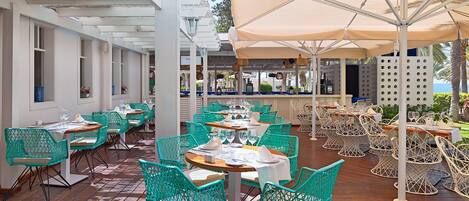 Un restaurante con terraza, con sillas turquesas y mesas blancas, bajo un paraguas grande.