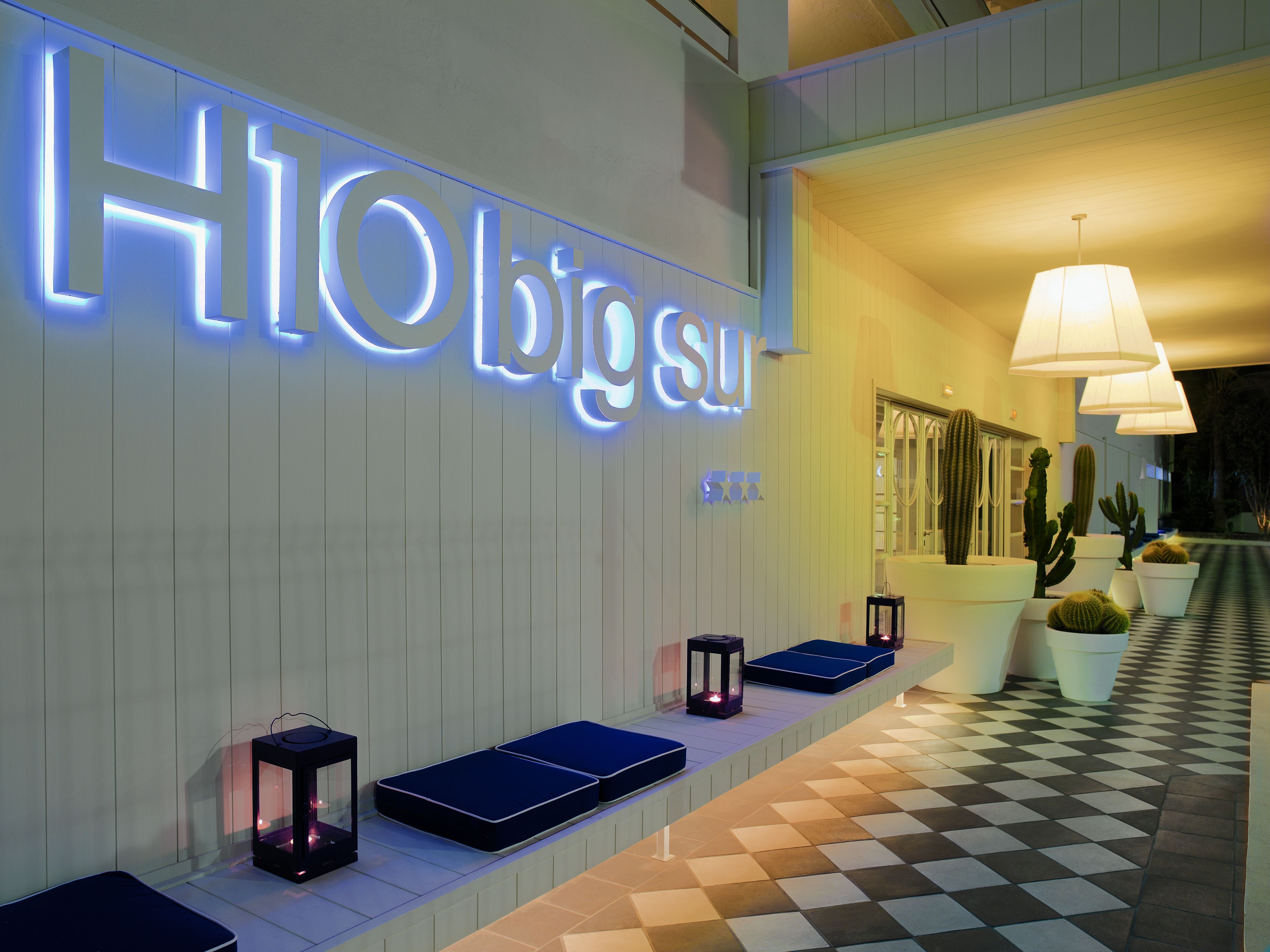 A hotel lobby with a neon sign reading "H10 big sur," a checkered floor, and several potted cacti.