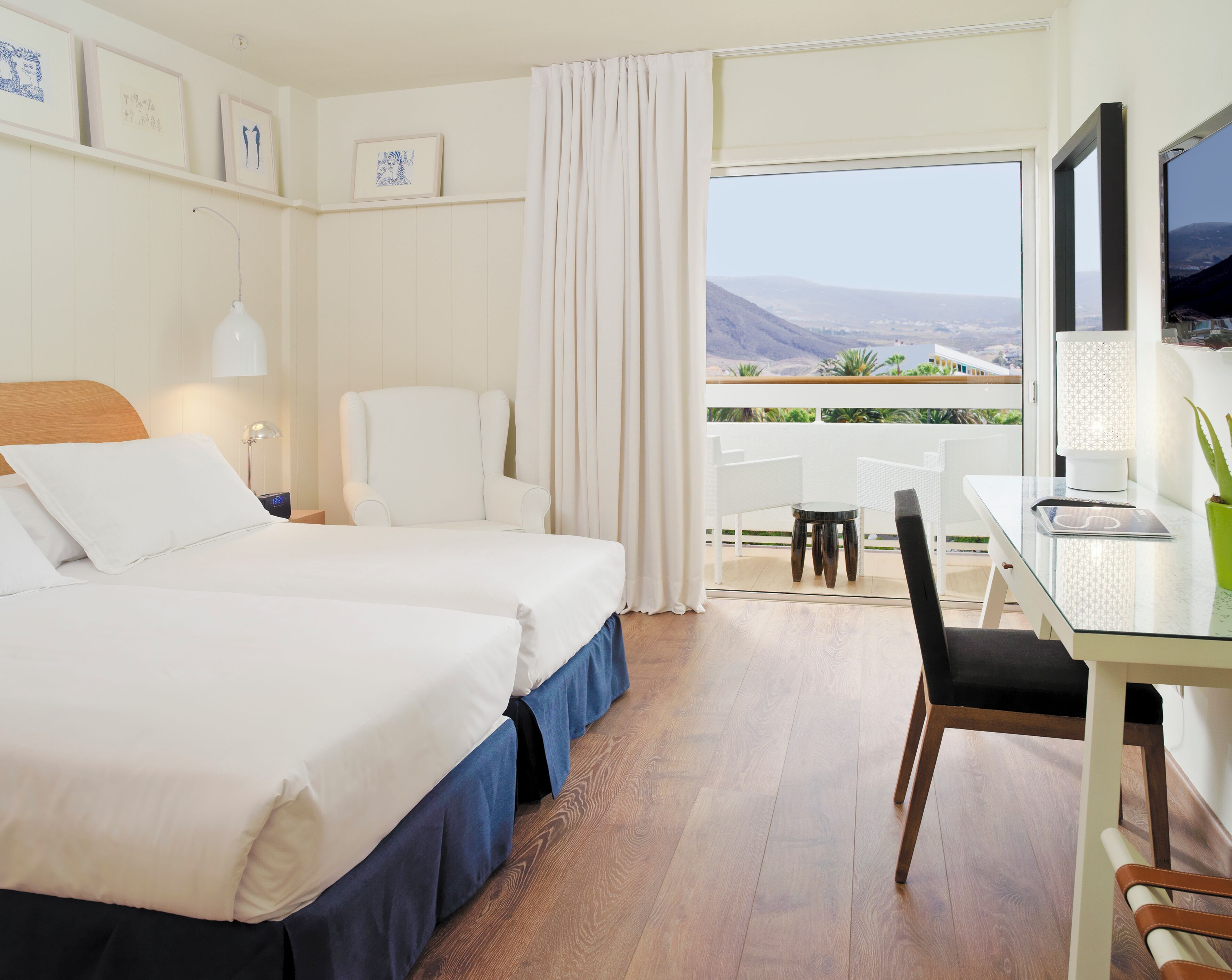 A hotel room with two beds, a desk with a chair, a TV, and a balcony with a view.